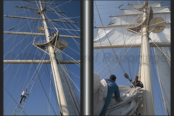 A sailor climbs the rope ladder that leads to the top of the fore mast while other sailors, reassemble the sail on the main mast